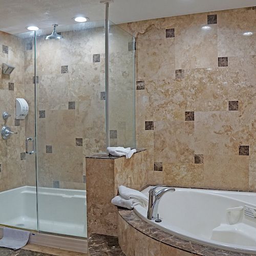 This image shows a bathroom with a glass-enclosed shower, a separate bathtub, and beige tiled walls. Towels are on the tub and shower ledge.