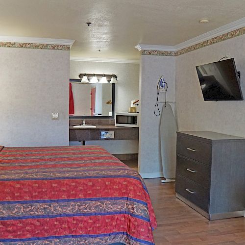 The image depicts a hotel room with a bed, TV, dresser, ironing board, refrigerator, and a bathroom sink area in the background.