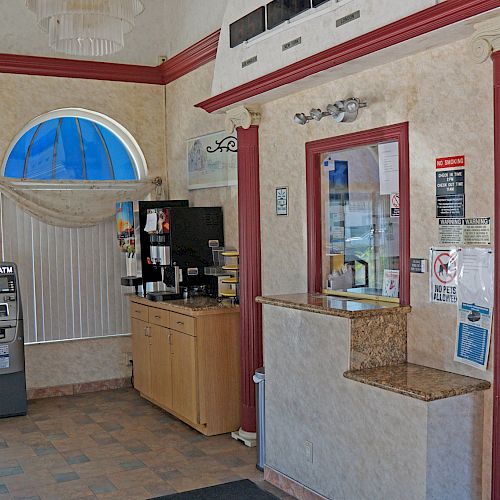 The image shows a small lobby area with a check-in counter, a coffee machine, an ATM, informational pamphlets, red chairs, and windows with blue awnings.