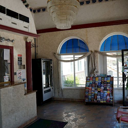 This image shows a small lobby area with an empty reception desk, a vending machine, brochures on a rack, red chairs, and arched windows.