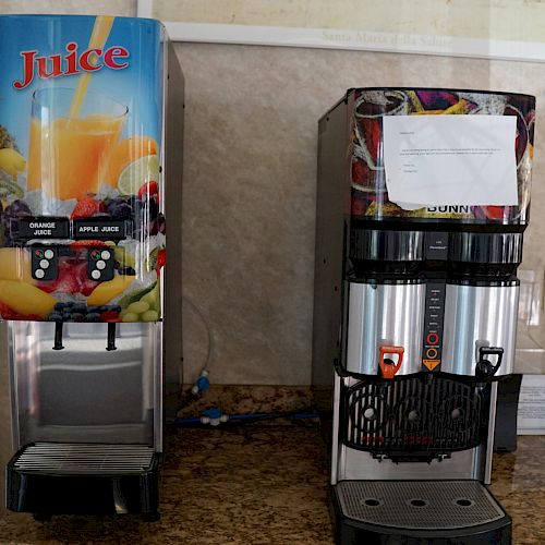 Two beverage dispensers are side by side on a counter; one for juice, the other with an unspecified label. On the right, stacked yellow plates.