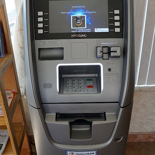 The image shows an Automated Teller Machine (ATM) with a screen, keypad, card slot, and cash dispenser in a room with a bookshelf and blinds.