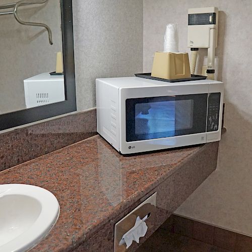 A microwave, coffee maker with cups, and sink are on a brown countertop in a room with a mirror and wall hook visible.