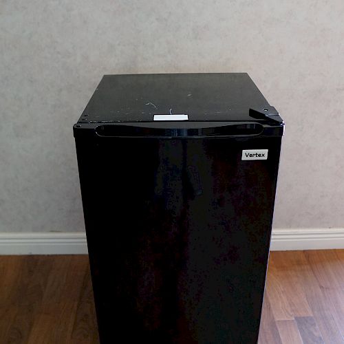 A black mini refrigerator stands on a wooden floor against a light-colored wall, with a small label on the front door.