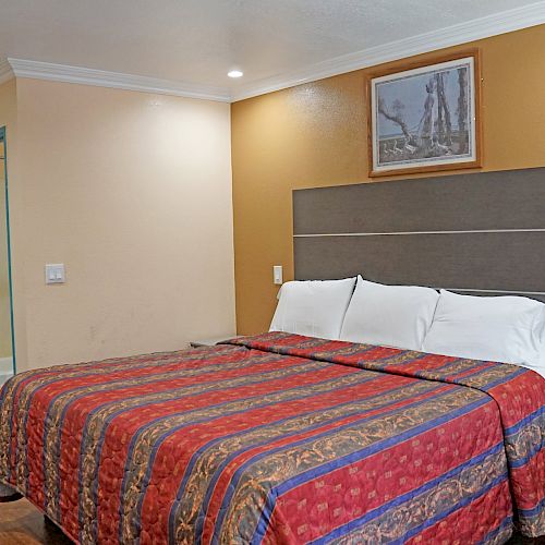 A hotel room features a bed with a colorful bedspread, a bedside table, a painting above the bed, and a visible bathroom area with a sink.