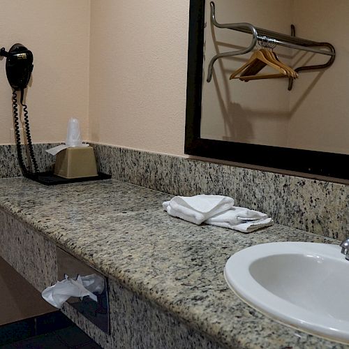 A bathroom countertop with a sink, soap, folded towels, a hairdryer, and a large mirror, with a mounted TV and clothes hangers in the reflection.