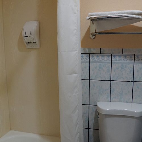 The image shows a bathroom with a bathtub, a shower curtain, a toilet, a soap dispenser, tiled wall, and a shelf with folded towels above the toilet.