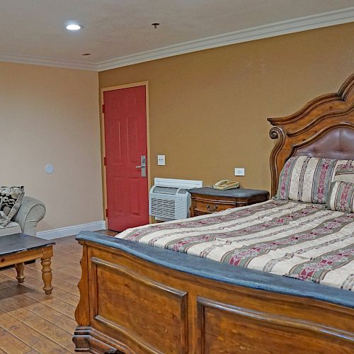 The image shows a bedroom with a wooden bed, a striped comforter and pillows, a couch, a coffee table, and a red door with a beige wall background.