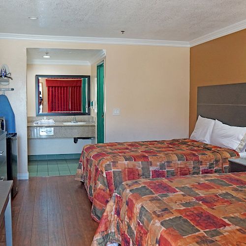 The image shows a hotel room with two beds, a flat-screen TV, a desk, an iron with an ironing board, and a sink area visible through an open doorway.