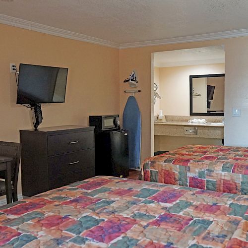 A hotel room with two beds, a wall-mounted TV, a small table with chairs, a dresser, a microwave, a mini fridge, an ironing board, and a bathroom.