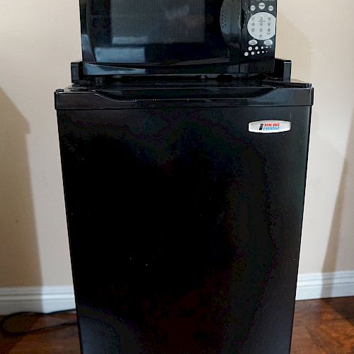 The image shows a black microwave oven placed on top of a black mini refrigerator against a beige wall on a wooden floor.