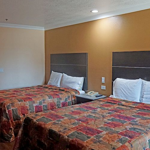 The image shows a hotel room with two double beds, colorful bedspreads, a nightstand, and an open view into a bathroom area with a sink.