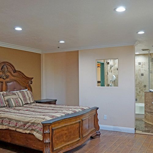 A bedroom with a large ornate bed, matching furniture, and an open bathroom featuring a whirlpool tub, under warm lighting.