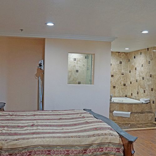 The image shows a spacious, well-lit bedroom with a bed, wooden dresser, and en-suite bathroom featuring a jacuzzi tub and tiled walls.