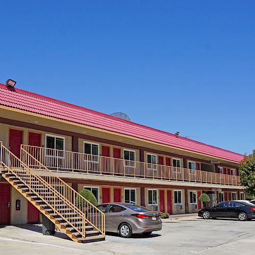 This image shows a two-story motel with red roofs, an outdoor staircase, and a parking lot with several cars parked under a clear blue sky.