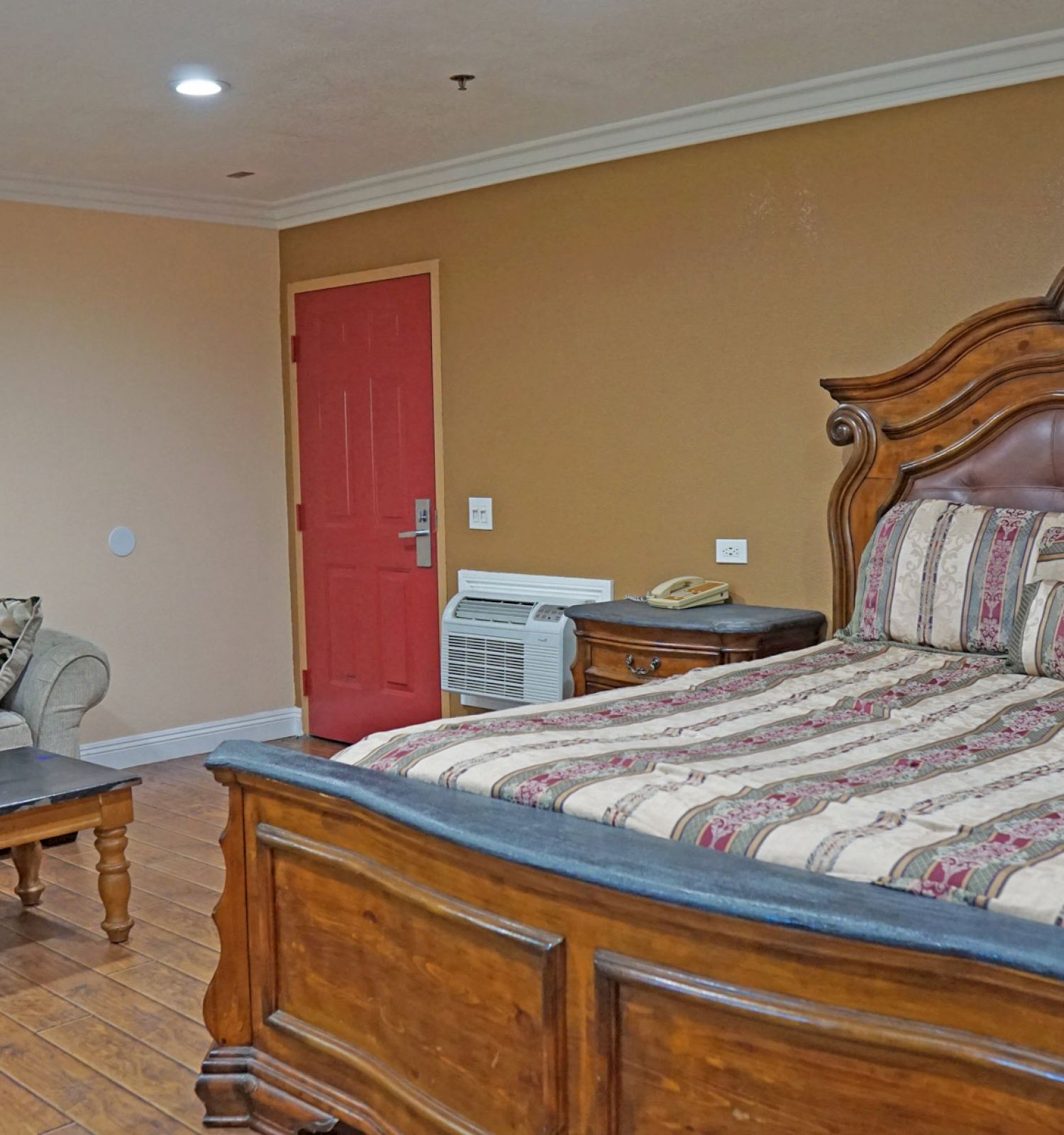 The image shows a cozy room with a large wooden bed, a patterned comforter, a beige couch, a wooden coffee table, and a red door in the background.