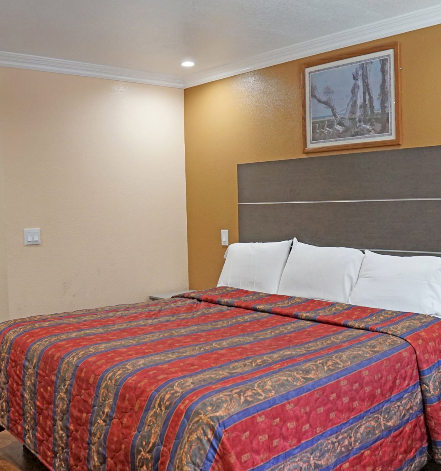 This image shows a hotel room with a neatly made bed, a bedside table, a lamp, and a painting on the wall. The room has warm colors and a sink area.