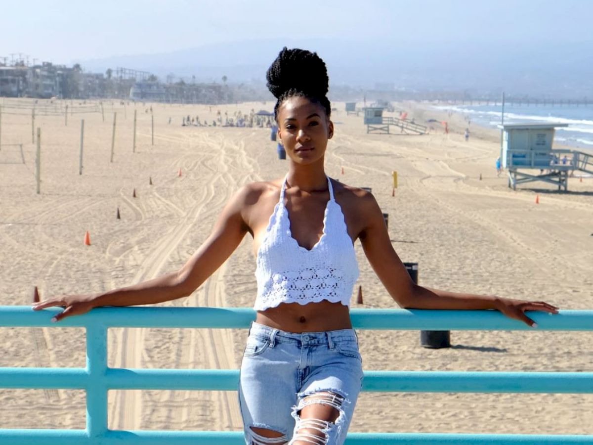 A woman leans against a turquoise rail on a pier, with a sandy beach, lifeguard tower, ocean, and beachside houses in the background.