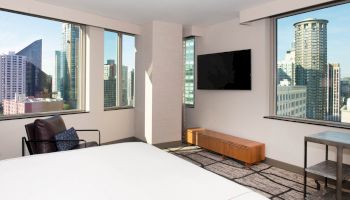 A modern hotel room with a bed, a chair, a TV, and large windows offering a cityscape view with tall buildings.