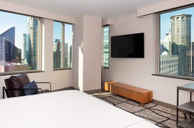 A modern hotel room with a bed, a chair, a TV, and large windows offering a cityscape view with tall buildings.