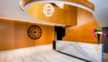 A modern lobby with wooden walls, a large wall clock, marble reception desk, decorative lighting, and a vase with colorful flowers.