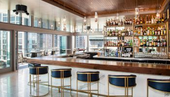 The image shows a modern bar with a wooden counter, stylish bar stools, and a well-stocked liquor shelf, in a bright space with large windows.