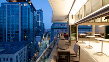A modern balcony with outdoor seating overlooks a cityscape filled with tall buildings. The sky is transitioning to night, adding a serene atmosphere.