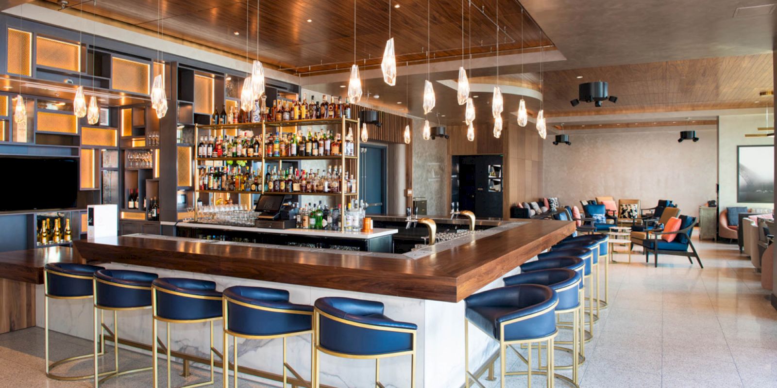 This image shows a modern bar with a large selection of bottles, stylish bar stools, hanging lights, and a spacious, elegant seating area.