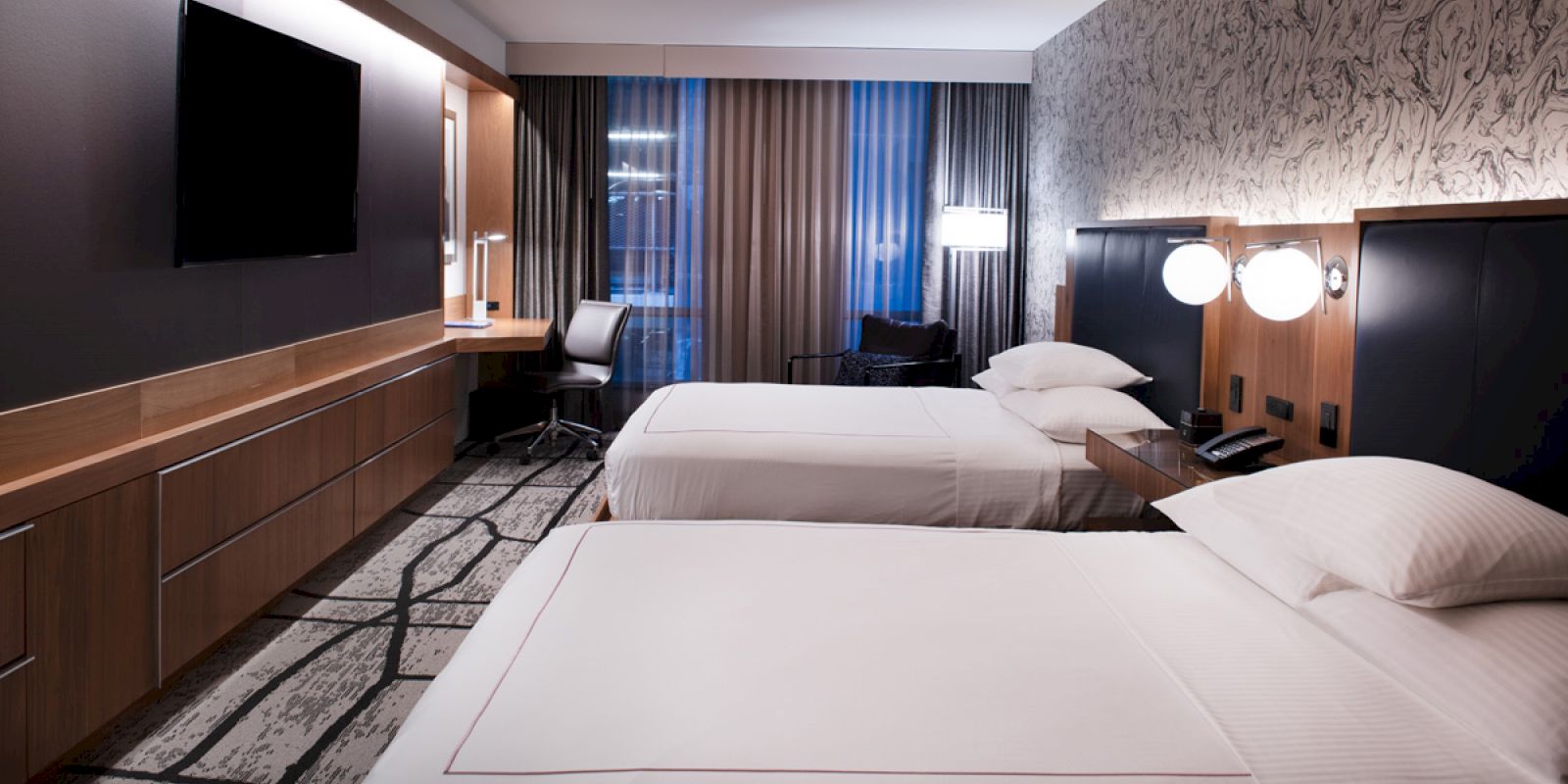A modern hotel room features two beds, a desk with a chair, a wall-mounted TV, and stylish decor with contrasting textures and patterns.