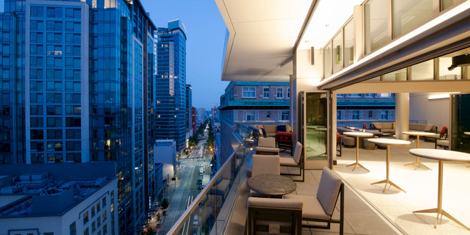 The image shows a modern cityscape from a high balcony with chairs and tables, overlooking tall buildings and a busy street below at dusk.