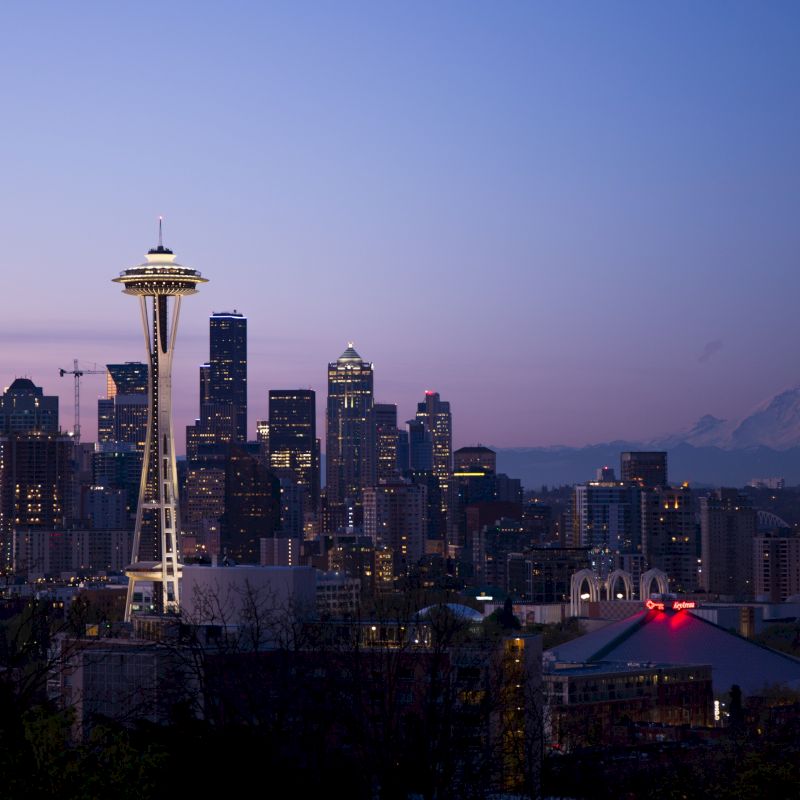 The image shows a city skyline during twilight with the Space Needle prominently featured and a mountain in the background against a clear sky.