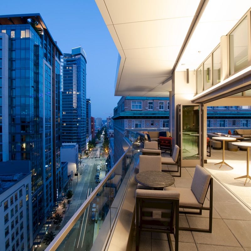 A cityscape view from a modern balcony at dusk, featuring tall buildings, a street below, and outdoor seating on the terrace.