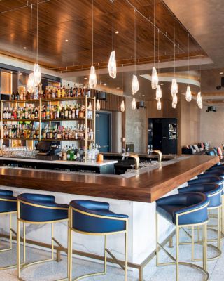 The image shows a modern bar with stylish blue stools, a well-stocked bar, cozy seating areas, and pendant lighting creating an inviting atmosphere.
