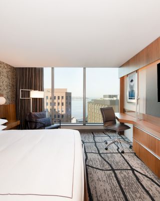 A modern hotel room features a large bed, wall-mounted TV, desk, chair, and a window with an urban view, and a patterned accent wall ends the sentence.