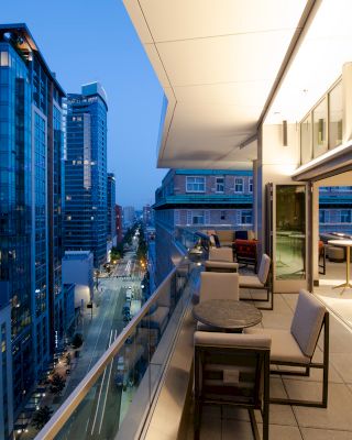 A modern, open balcony overlooking a city street with high-rise buildings at dusk. The balcony has tables, chairs, and seating areas.