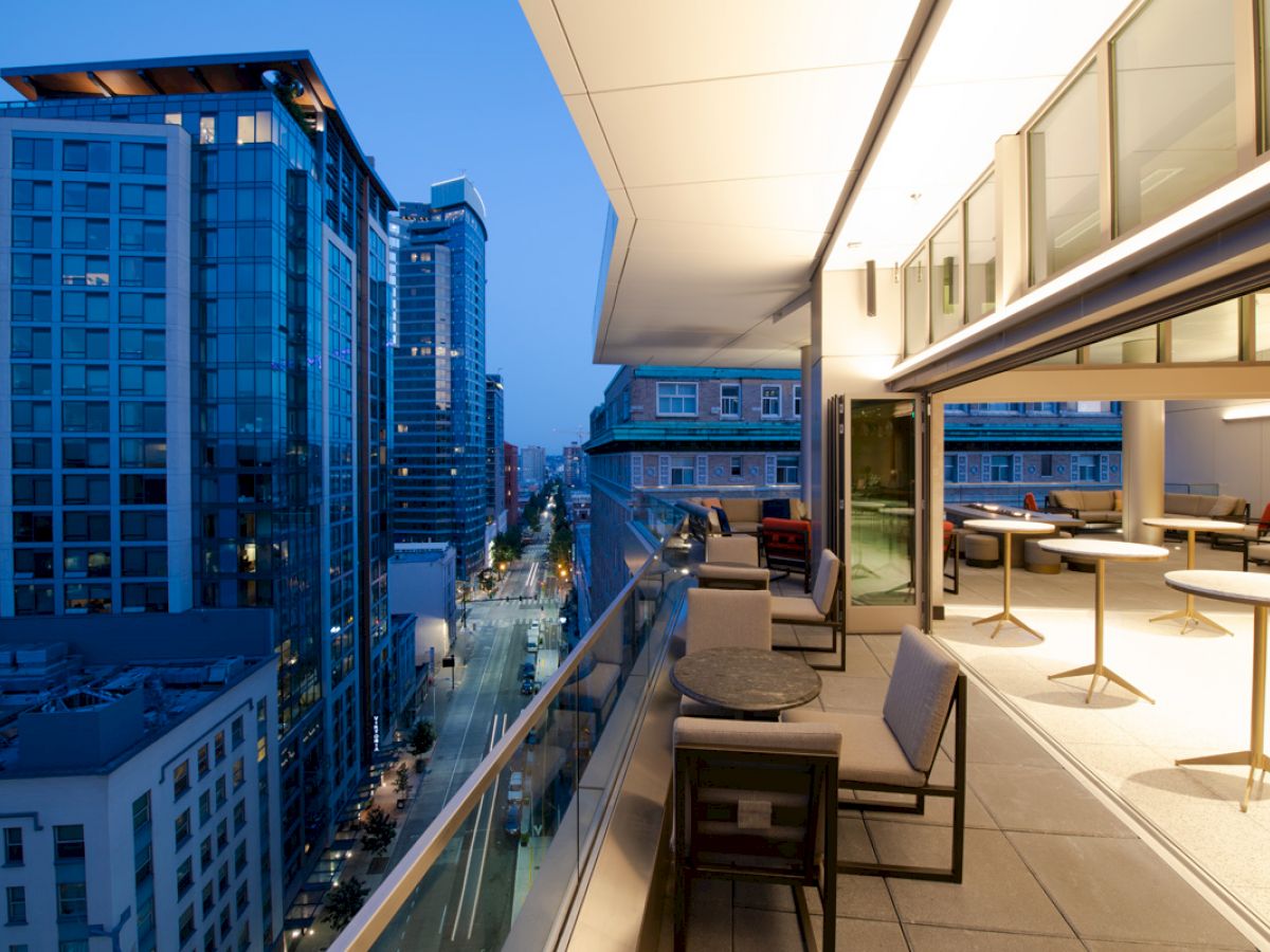 This image shows a modern balcony with seating, overlooking a cityscape at dusk, with high-rise buildings and a well-lit extended area.
