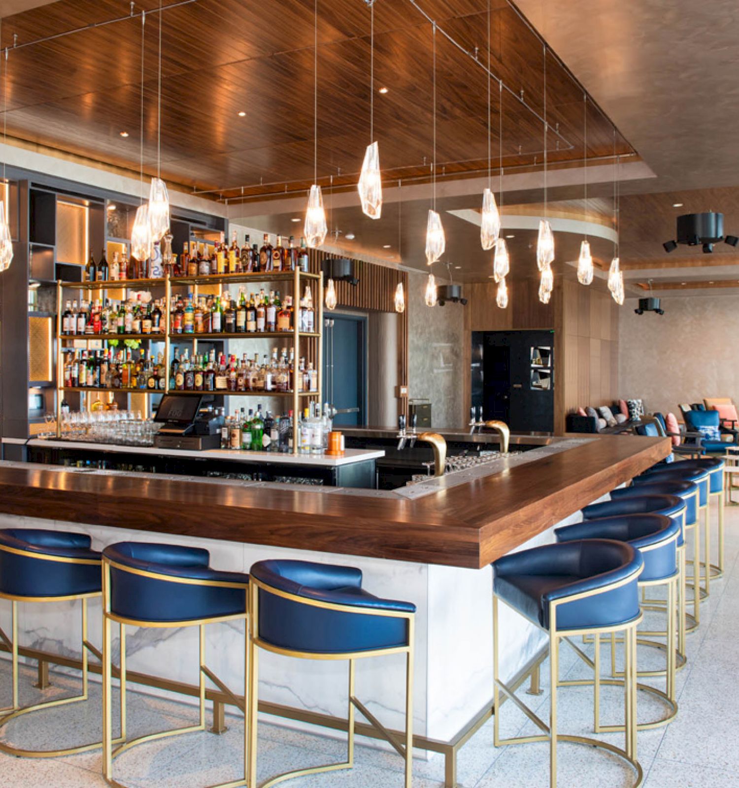 A modern bar with a central counter, blue barstools, a variety of bottles, and hanging light fixtures, with lounge seating in the background.