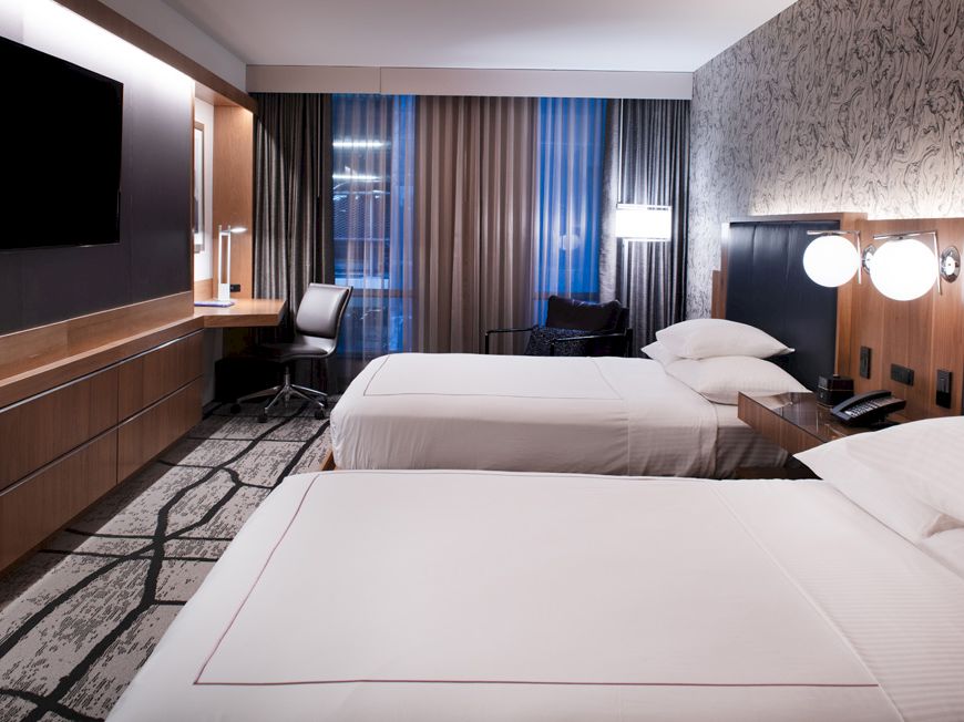 A modern hotel room with two beds, a desk, a flat-screen TV, and large windows with curtains. The room has contemporary decor and lighting.