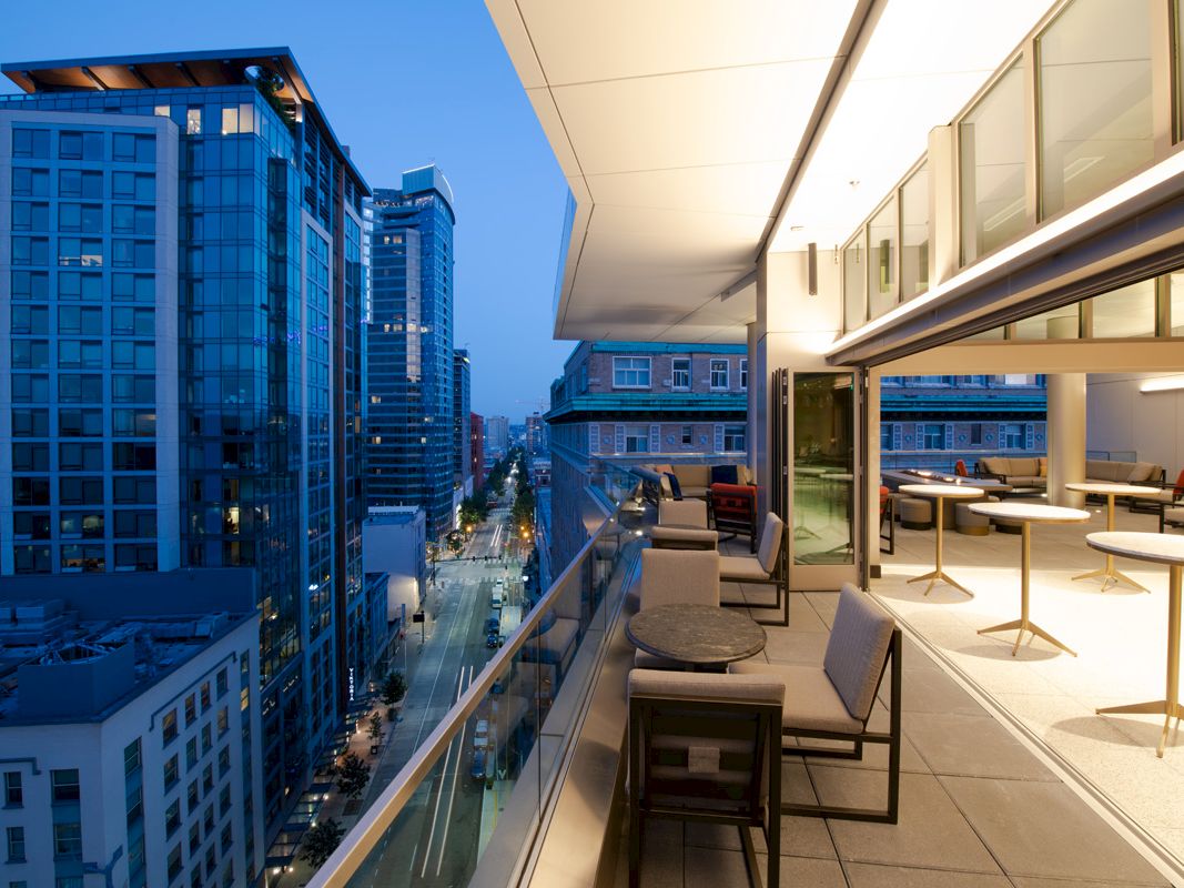 A modern balcony with tables and chairs overlooks a city street lined with tall buildings at dusk.