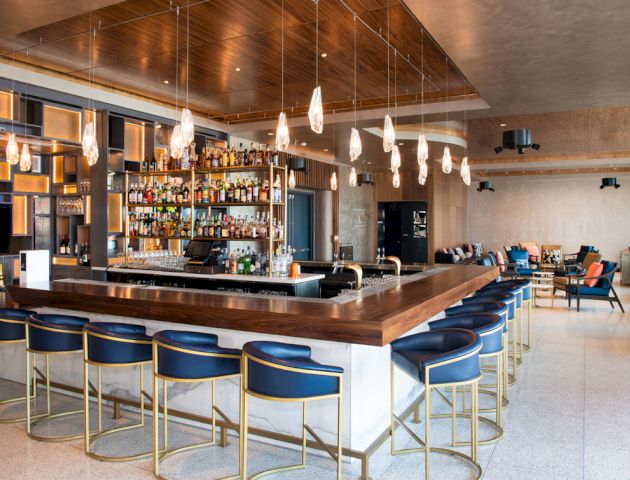 The image shows a modern bar with wooden and metallic accents, a well-stocked liquor shelf, bar stools, pendant lights, and a seating area.