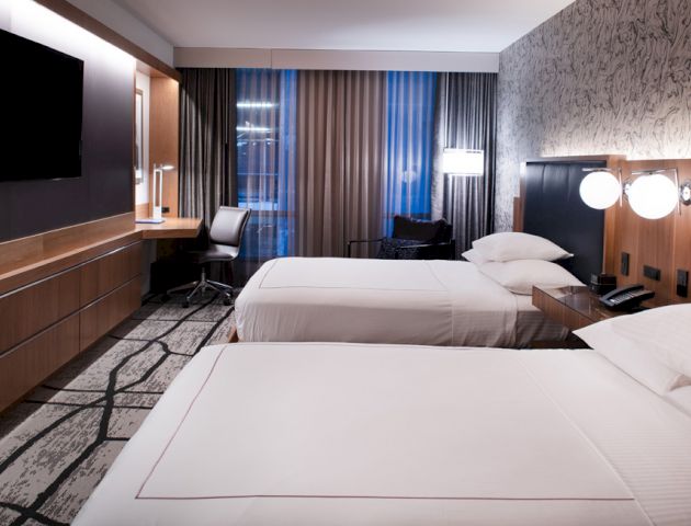 The image shows a modern hotel room with two neatly made beds, a desk with a chair, a wall-mounted TV, and stylish decor.
