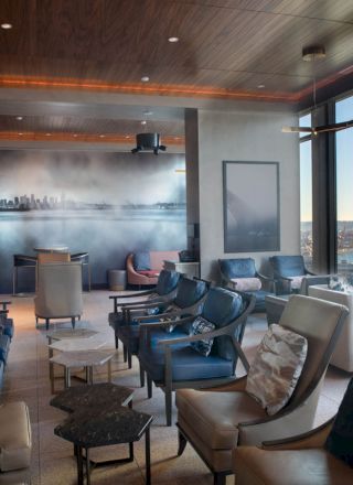 A modern lounge area with comfortable seating, large windows offering a view of a waterbody and skyline, and a wall mural of a cityscape.