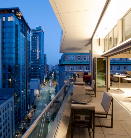 The image shows a modern cityscape at dusk with tall buildings and a balcony area furnished with chairs and tables, overlooking a street below.