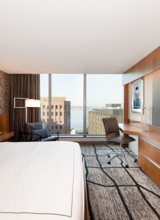 A modern hotel room with a large bed, a desk, chair, wall-mounted TV, and a window with a cityscape view.