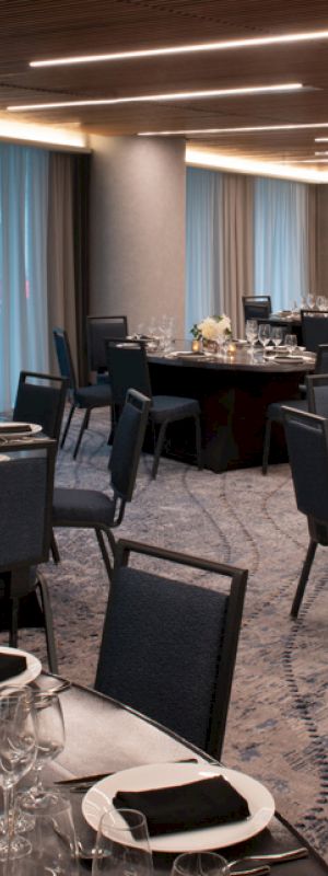 The image shows a banquet room with round tables set for dining, each with glassware, black napkins, and floral centerpieces, under modern lighting.