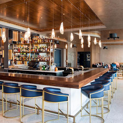 The image shows a stylish bar with a wooden counter, blue chairs, pendant lights, and a fully stocked bar. The lounge area is in the background.