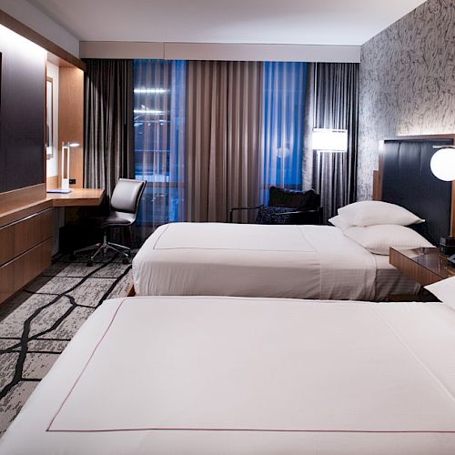 The image shows a modern hotel room with two beds, a TV, a desk with a chair, and a well-lit design featuring a patterned rug and wall.