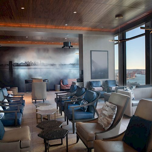 A modern lounge with ample seating, a wall mural, large windows showcasing a waterfront view, and a ferris wheel visible in the background.