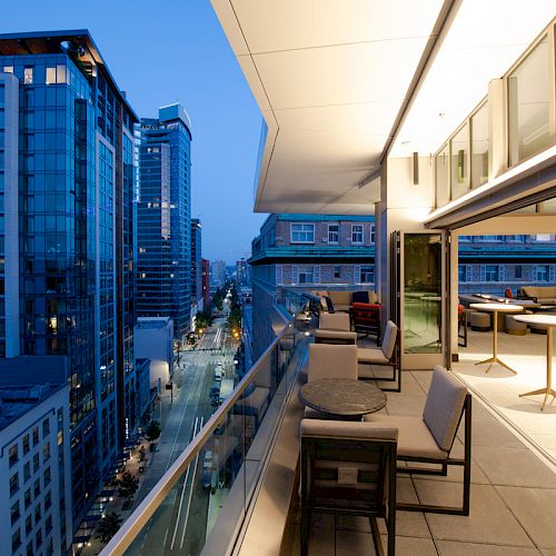 This image shows a modern cityscape at dusk with tall buildings, along with a stylish terrace featuring tables and chairs.