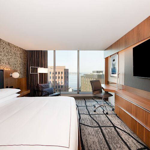 A modern hotel room with a large bed, flat-screen TV, desk, chair, and panoramic window showing a cityscape and waterfront view.